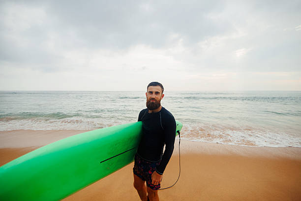 istock Young  bearded man with surfboard standing near a beach. Man with surfing board outdoors on a summer day. 2236893525