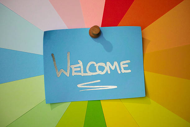 The single word 'WELCOME' written in a silver marker pen on blue note paper, pinned to a colorful array of background blank message notes that radiate around the main message note in a rainbow spiral effect. Conceptual greeting message of welcome, welcome sign, etc.
