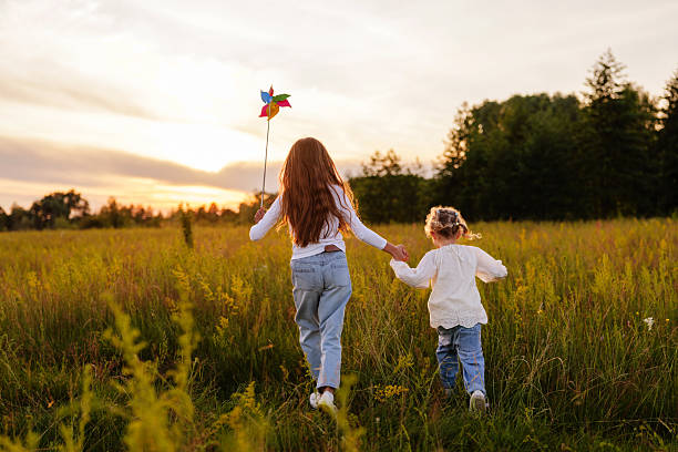 istock Two sisters run away from the camera through a sunny field at sunset, holding hands as the older girl leads the way with a colorful pinwheel. 2236909578