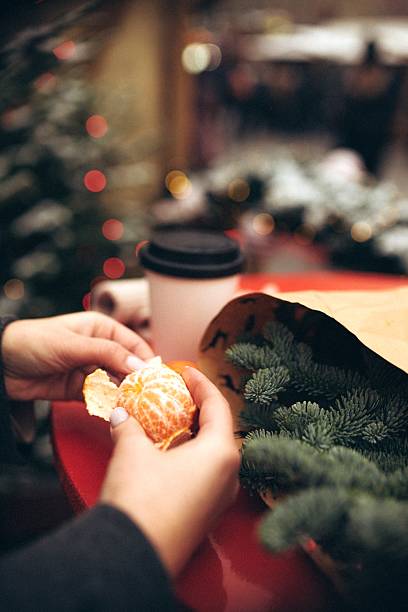Hands peel a mandarin on a bright red table, with festive greenery and a warm beverage nearby. Soft lights create a cozy atmosphere, perfect for enjoying seasonal treats.