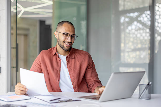 istock A happy man in glasses reviews documents and uses his laptop in a bright, modern office setting. 2236912464