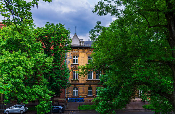 Krakow, Poland – May 22, 2025: A yellow historic townhouse partly hidden behind lush green trees creates a picturesque urban view. Classic architecture blends with natural surroundings in the Old Town district.