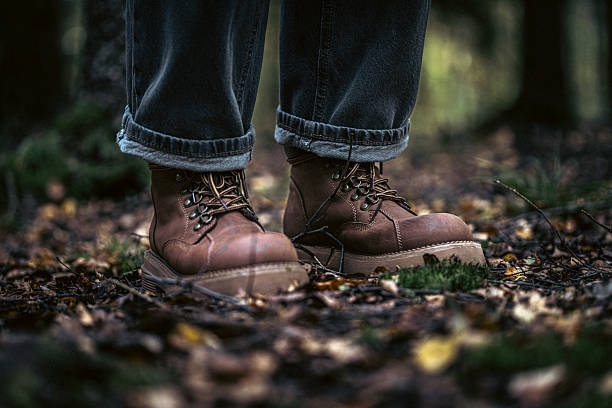 Close-up of sturdy brown boots resting on a leaf-strewn forest floor. Jeans cuffed at the ankle, suggesting outdoor exploration, adventure, and rugged outdoor lifestyle in a peaceful natural setting.