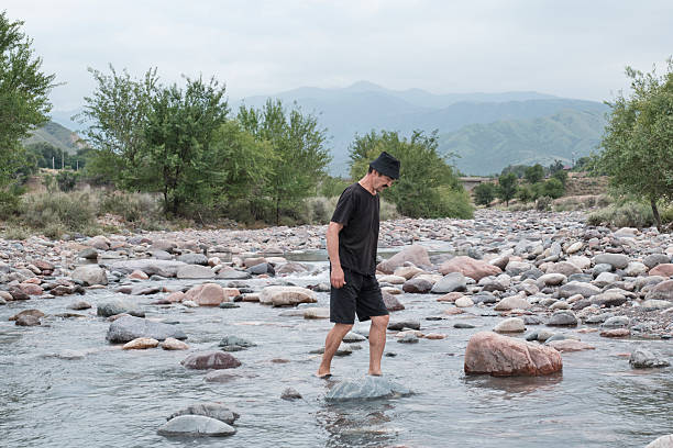 A senior man in black clothes and a Panama hat crossing a shallow rocky mountain river, surrounded by green trees, on a cloudy day. Elderly people, lifestyle concept