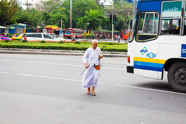 Female thai mok in white clothes is crossig street Phahoyotnin Road at rounabout at Victory Monumeny. She has exited a bus and is walking around a standing bus. View from sidewalk and  bus top onto street. Woman is a senior woman. In background is traffic. Victory Monument is one of main bus stops in Bangkok at roundabout of Phahonyothin Road in Phaya Thai