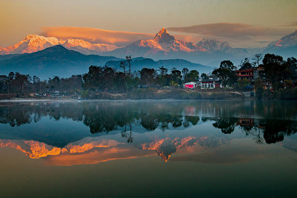The majestic Annapurna range glows in golden hues at sunrise, with Machapuchare’s iconic peak rising above the clouds. The calm waters of Phewa Lake perfectly mirror the snow-capped Himalayas, creating a breathtaking reflection of nature’s grandeur. Nestled below, the lakeside settlement blends harmoniously with the serene landscape. This scene captures the awe-inspiring beauty of Nepal’s mountains, where light, water, and sky meet in perfect balance.