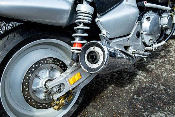 Close-up of a motorcycle rear wheel and exhaust system displaying chrome and gray features in a garage environment, sunlight illuminating the metallic surfaces.