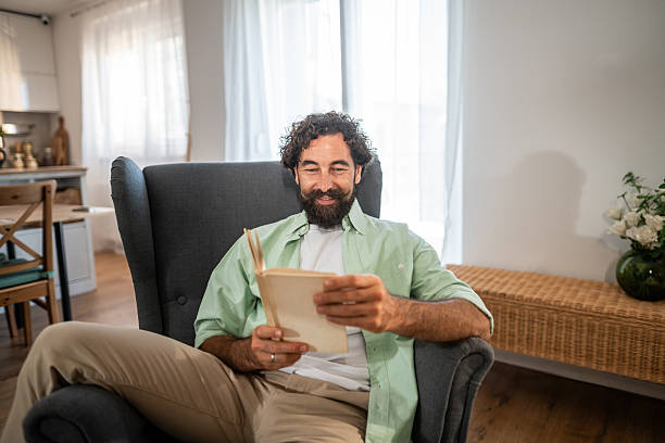 istock Man smiling while relaxing and reading book in armchair at home 2236937740