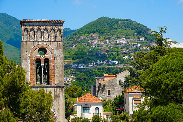 Historic bell tower in Ravello, Italy, with the terraced hillsides of the Amalfi Coast in the background. The traditional Mediterranean houses, vineyards, and lush greenery create a picturesque and authentic Italian landscape.