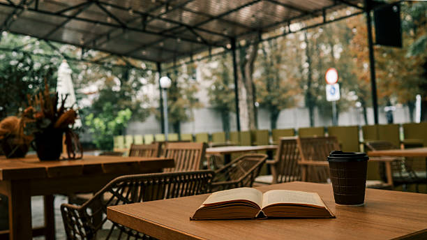 An open book and a cup of coffee sit on a wooden table in an outdoor cafe setting, creating a relaxing atmosphere.