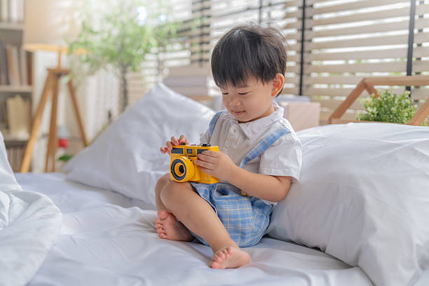 Cute Asian child boy plays with toy camera while resting on white soft bed near big window during weekend morning, capturing precious happy childhood lifestyle moments at home