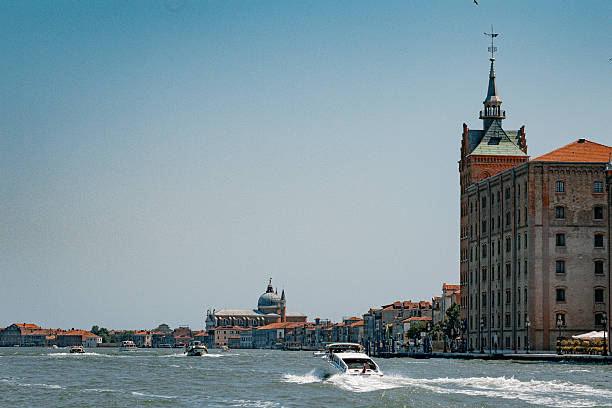 This photo captures a dynamic view of Venice, Italy, taken from the lagoon. In the foreground, a motorboat speeds across the shimmering water, leaving a white trail of waves behind. To the right stands a tall historic building with a pointed spire, while in the distance, the domes of Venetian churches rise gracefully above the terracotta rooftops. The clear sky adds to the bright and lively atmosphere of the scene.