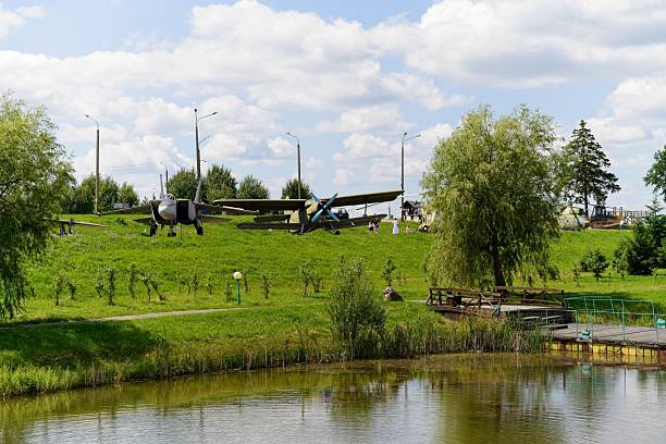 Minsk region, Belarus, July 12, 2025.Vintage aircraft are displayed on a grassy hill in the Minsk region, Belarus, part of the Stalin Line historical and cultural complex. The scene includes several trees and lamp posts surrounding the aircraft, set against a backdrop of a clear, partly cloudy sky. In the foreground, a small pond reflects the landscape, bordered by wooden walkways. The Stalin Line is a historical defense line, and the site may serve as a museum or memorial area showcasing military history.