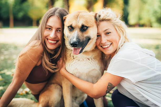 Happy moment with two young women and their dog, hugging and smiling together in the park. Concept of friendship, fun, summer lifestyle, and pet love