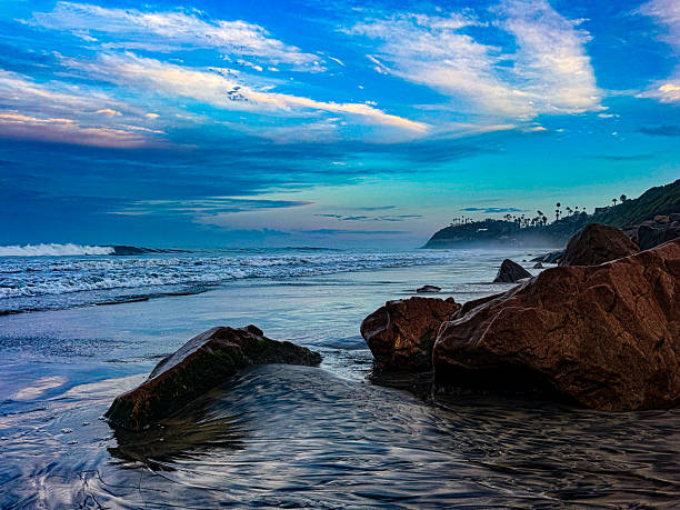 This is a beautiful sunrise photo taken early in the morning at Swamis beach California with rocks in the colorful magenta orange sand reflection of the sun as the waves lap against the stony shore.