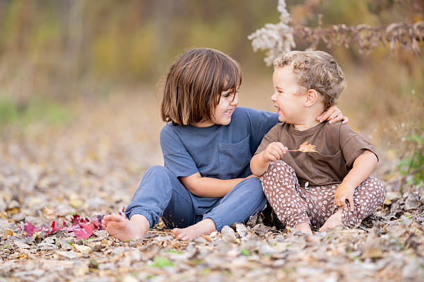 Two children sit barefoot on fallen autumn leaves, sharing a joyful moment as they look at each other and smile. A candid outdoor scene conveying friendship, play, and seasonal warmth.