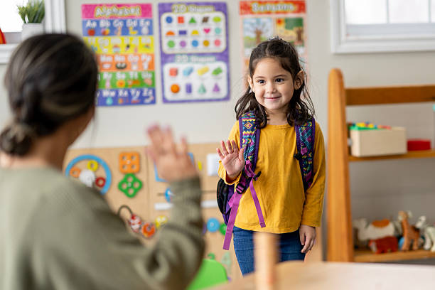istock Front-Facing Hispanic Girl With Backpack Waves Hello To Teacher In Colorful Classroom Setting 2236996191