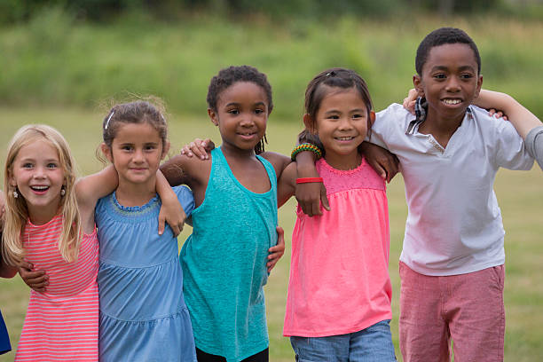 A multiethnic group of young children stand side by side in a sunny open field, smiling and linking arms, showcasing friendship, teamwork, and outdoor fun among friends.
