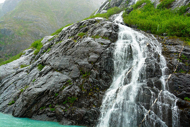 A strong and steady waterfall flows over a dark rocky cliff into turquoise glacial waters. Photographed in Alaska, the image captures the raw power and refreshing energy of natural mountain runoff.