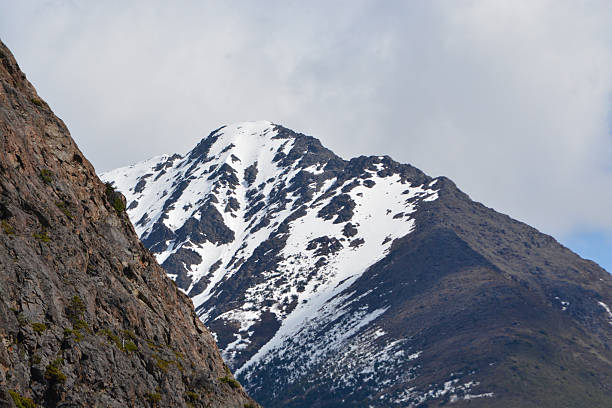 A dramatic view of a rugged mountain ridge with snowy summit and rocky foreground, photographed in Alaska. The contrast between terrain types makes this image ideal for geological, adventure, or environmental themes.