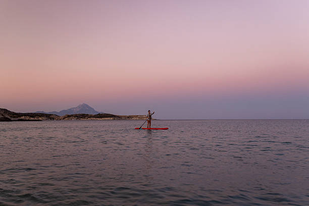 A lone individual enjoys paddleboarding on calm waters during a serene sunset, with a pastel-hued sky and distant mountains enhancing the peaceful scene.