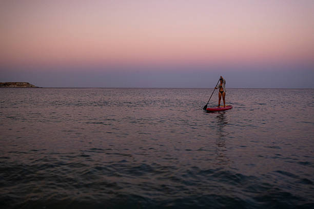 A woman paddleboards on a serene ocean at sunset. The pastel-colored sky creates a tranquil atmosphere, blending soft pink and purple hues with calm waters.