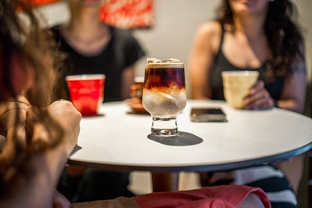 Young professionals gather at white cafe table enjoying layered iced coffee drinks in casual modern setting with natural lighting.