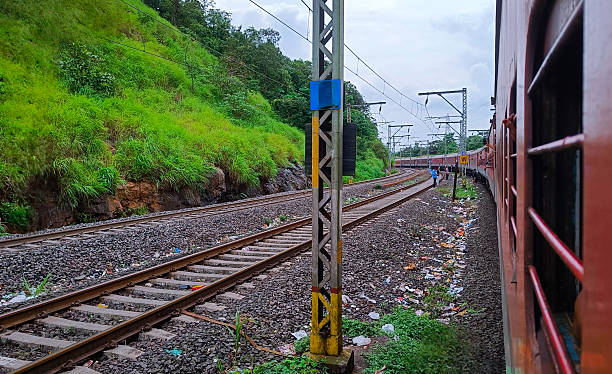 This image captures the view from a moving train in India, showing a long, red passenger train curving along the tracks. The railway is flanked by a lush, green hillside on one side and littered ground on the other, under an overcast sky. The perspective highlights the parallel lines of the tracks leading into the distance, conveying a sense of travel and movement through a scenic, yet realistic, landscape.