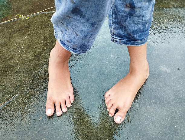 This image captures a low-angle view of a person's bare feet standing on a wet, dark-colored pavement. The person is wearing faded and stained blue denim shorts, and their feet and toenails show signs of being outdoors.