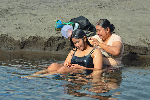 A teenage girl smiles softly as water flows across her face while her mother rinses her hair with a plastic container, both sitting calmly in a shallow seaside creek.
