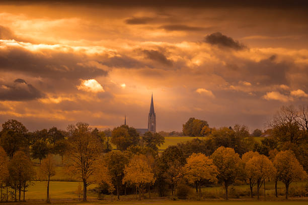 A sky full of heavy clouds dips the scenery in a warm, orange light. In the center is a church with a high church tower, surrounded by autumn-colored trees, while a flock of birds circles over the top.