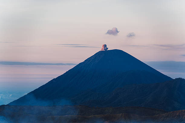 A volcano Mount Semeru emitting smoke against a pastel sky during sunrise