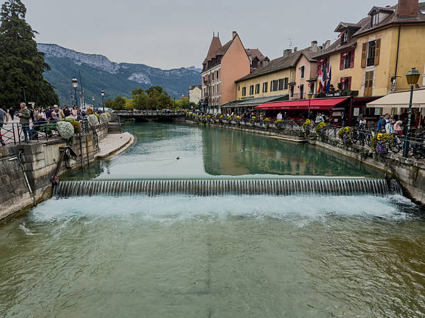 A river runs through a town with a bridge over it. The water is clear and the bridge is in the middle of the river. There are many people walking around the town, some of them near the water