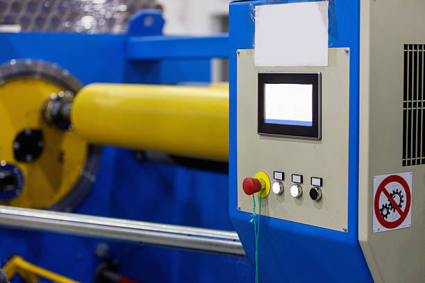 A close-up view of an industrial control panel on blue machinery featuring a digital screen, red emergency stop button, smaller white buttons, and a safety sign, highlighting manufacturing plant operations and safety protocols.