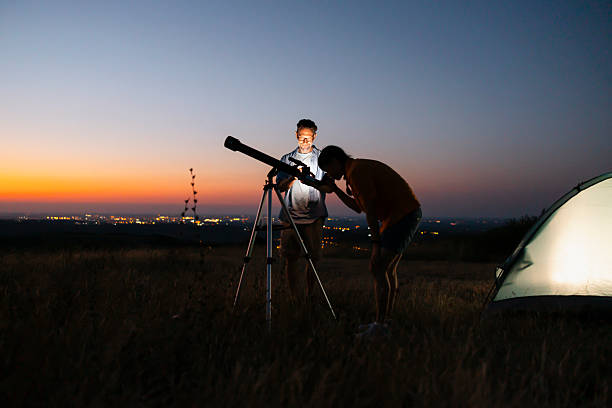 Mature multi-ethnic couple enjoying their leisure time outdoors, stargazing and watching stars using telescope and smart phone app while camping in nature at night