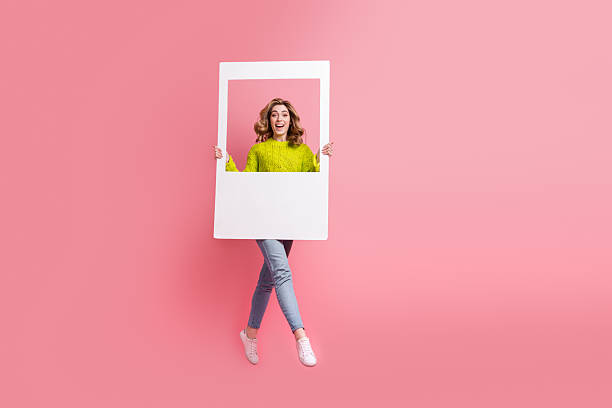 istock Adorable young woman with playful expression posing through a white frame on a pastel pink background wearing a yellow sweater 2237079187