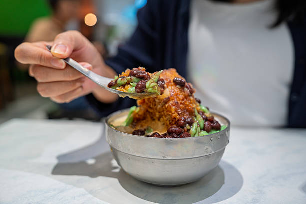 Close-up of a woman eating cendol, a traditional Southeast Asian dessert made with shaved ice, red beans, green rice flour jelly, and palm sugar syrup, served in a metal bowl.