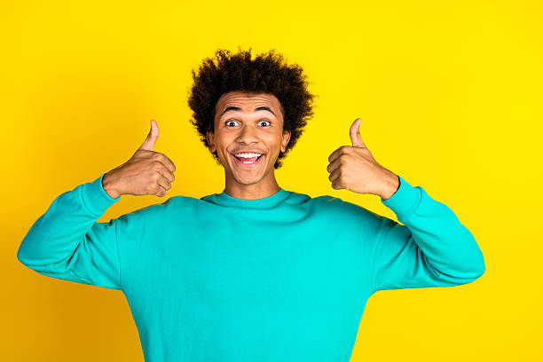 istock Cheerful young man in a vibrant teal sweatshirt showing thumbs up while smiling brightly against a yellow backdrop 2237080546