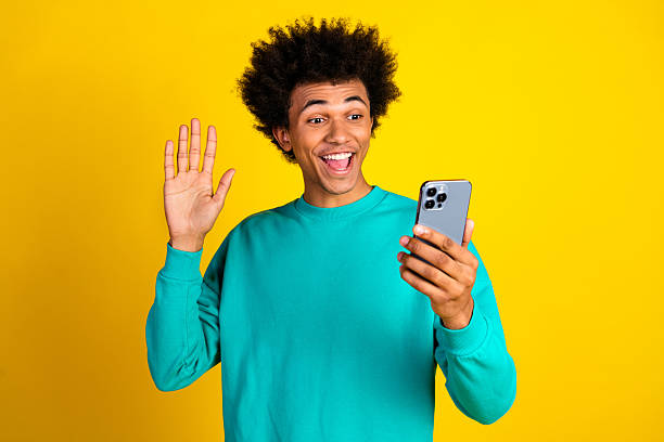 istock Cheerful young man in teal sweatshirt waving at smartphone while smiling against bright yellow background 2237081023