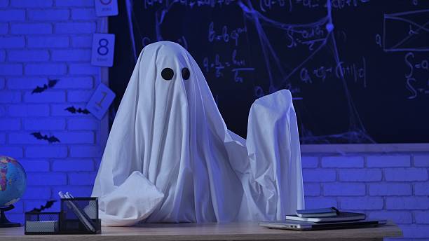 istock A person in a white ghost sheet sits at a table in a blue-lit room and points to the side. A monster teacher teaches in a classroom with a blackboard. Halloween concept. 2237082535