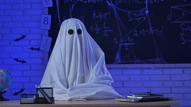 istock A person in a white ghost sheet sits at a table in a room with blue lighting. A monster teacher teaches in a classroom with a blackboard. Halloween concept. 2237082680