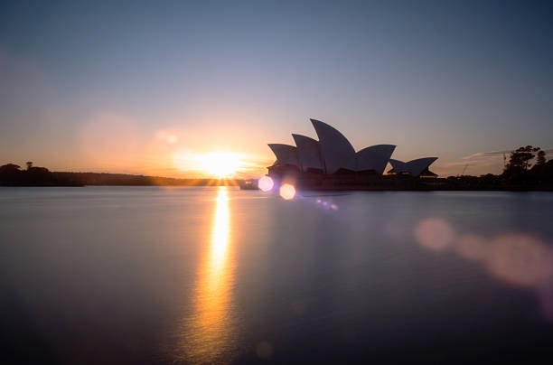 Sydney Australia - August 24, 2025: Early morning in Sydney, and the iconic Opera House and a glorious pre-dawn sky are reflected in the waters of Circular Quay.  A long exposure smooths out the water and genuine lens flare is created by the sun's positioning at the side of the iconic structure.