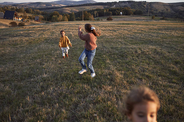 Portrait of three children playing game of catch, jumping and running on dry grass hay field paths in sunset. Trees and meadow on background. Looking around. Cloudy sunny sky. Haying time