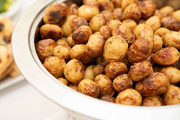 Close-up of a person serving seasoned roasted baby potatoes from a large outdoor grill onto a white disposable plate. The image captures the vibrant texture and color of grilled meat, potatoes, and other food items in a bustling street food setting. The scene conveys a warm, savory atmosphere and a sense of culinary craftsmanship.