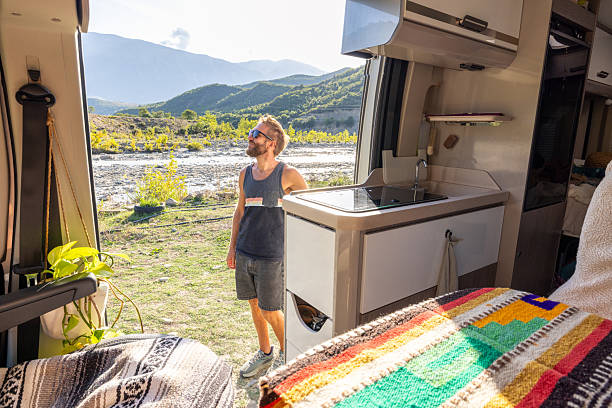 Bearded man in sunglasses standing outside camper van by river and mountains. Casual traveler appreciating outdoor scenery on a sunny day. Adventure and road trip lifestyle concept.