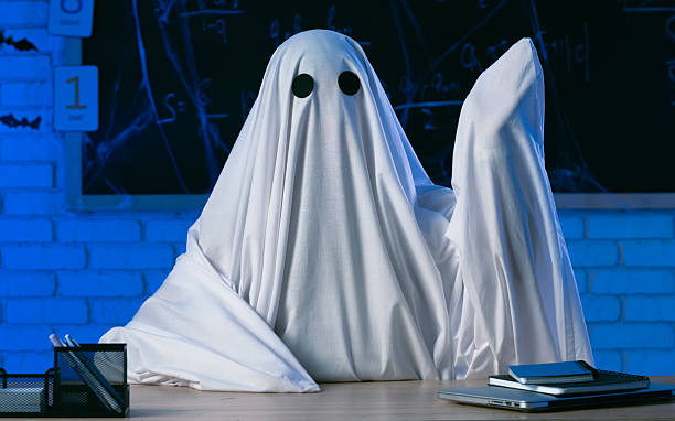istock A person in a white ghost sheet sits at a table in a blue-lit room and points to the side. A monster teacher teaches in a classroom with a blackboard. Halloween concept. 2237085414