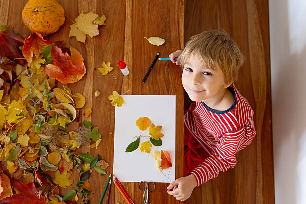 Sweet child, boy, applying leaves using glue while doing arts and crafts at home, autumntime