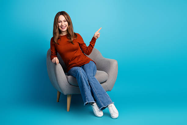 istock Attractive young woman pointing and smiling cheerfully while sitting on a modern chair against a blue background 2237091103