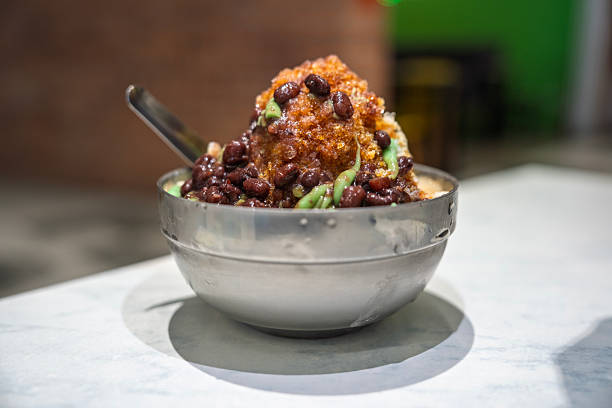 A bowl of cendol topped with red beans, palm sugar syrup, and shaved ice served in a metal bowl on a table.