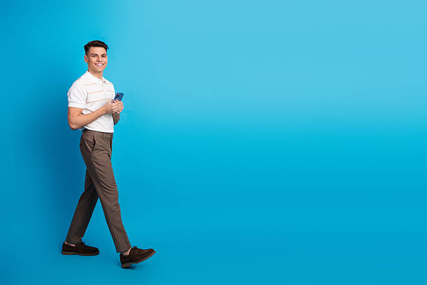 istock Young man walking while holding smartphone, dressed in casual attire, isolated against a vibrant blue background 2237091702